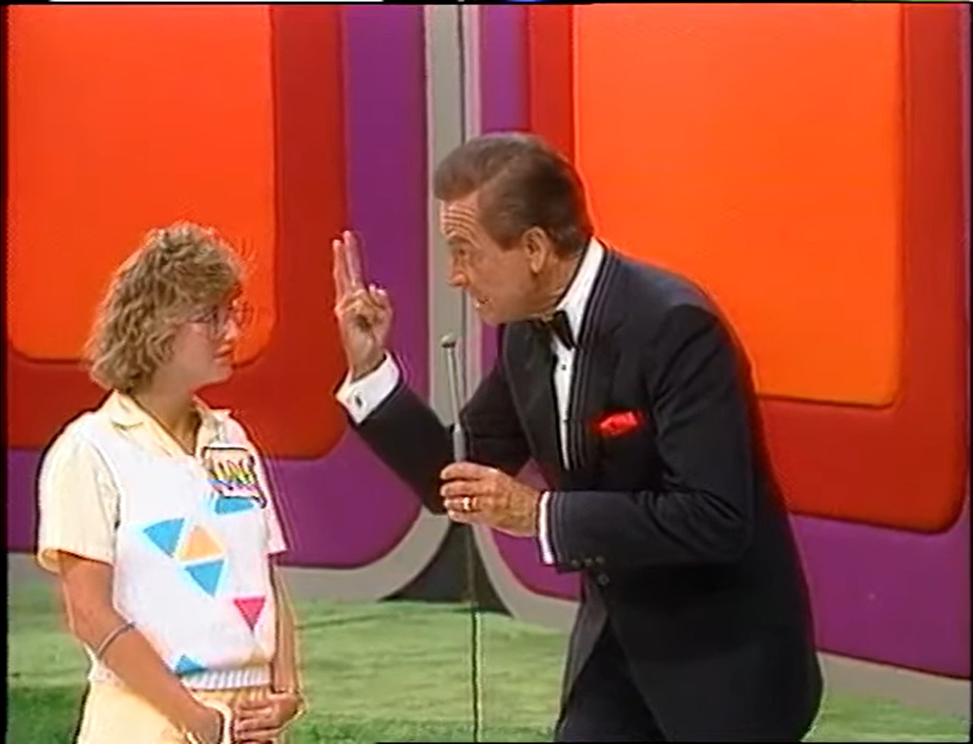 A game show host gives instructions to a female contestant during a game segment on The Price is Right, with colorful studio backgrounds.