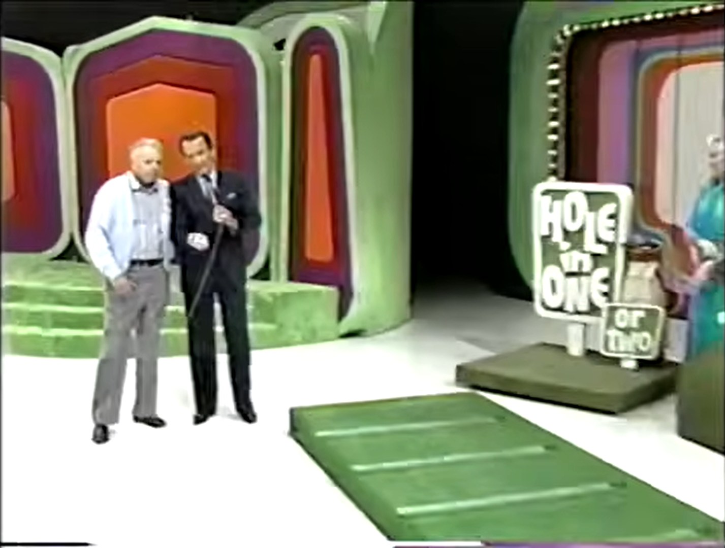 A contestants and a host stand on the set of Hole in One (or two) with a putting green and the game sign visible in the background.