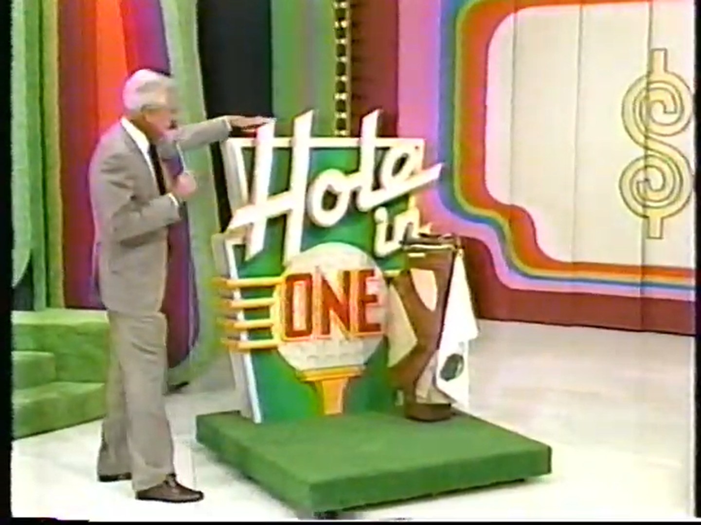 Bob Barker stands beside the colorful 'Hole in One' game sign on a game show set, showcasing the vibrant background and a putting green.