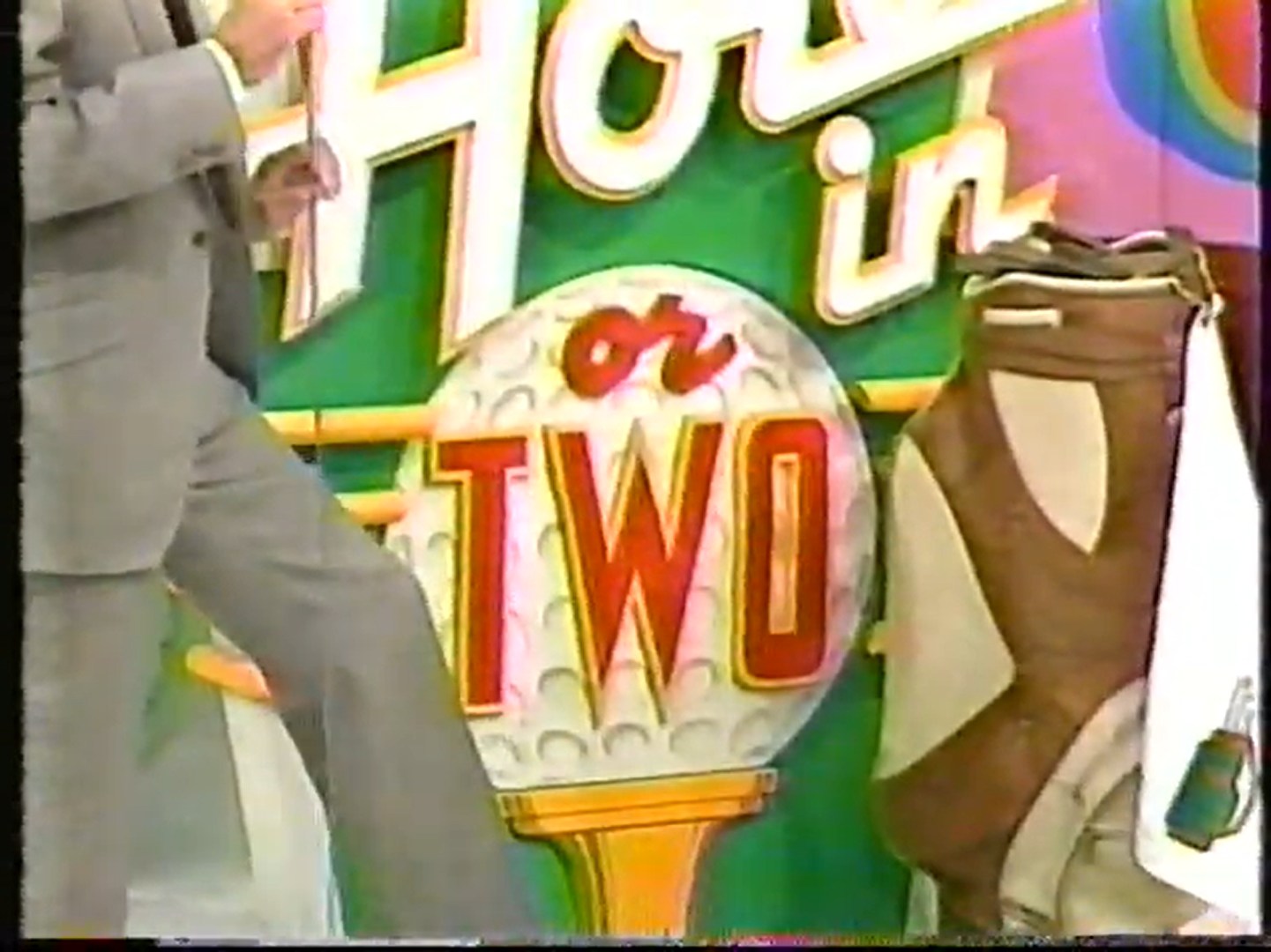 Bob Barker host standing next to a colorful backdrop featuring the words 'Hole in One or Two' with a golf ball design and a golf bag in the foreground.
