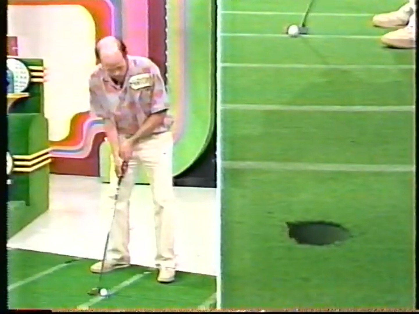 A contestant is preparing to putt a golf ball towards a hole on a putting green in a game show setting, with a splitscreen showing two angles of the putt.