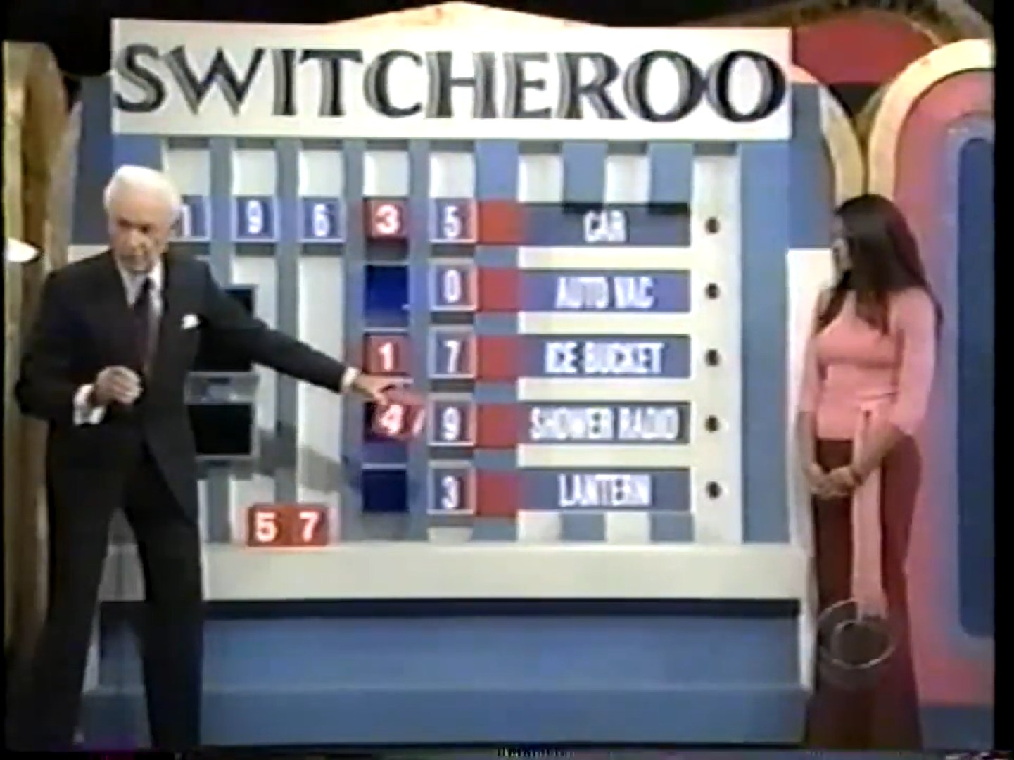 A game show host points to the Switcheroo game board displaying various prize options and missing digits, with a contestant standing nearby.
