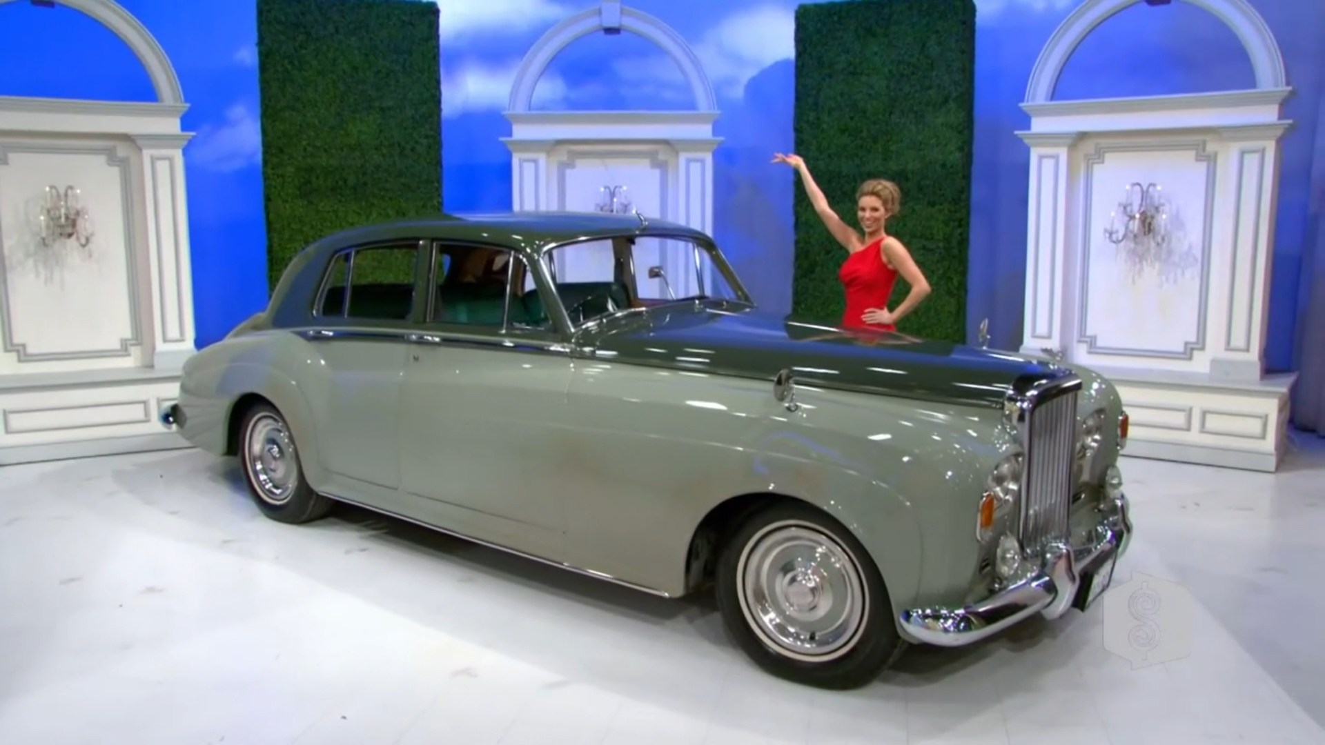 A woman in a red dress stands beside a classic gray Bentley, showcasing the car with a smile in a studio setting.