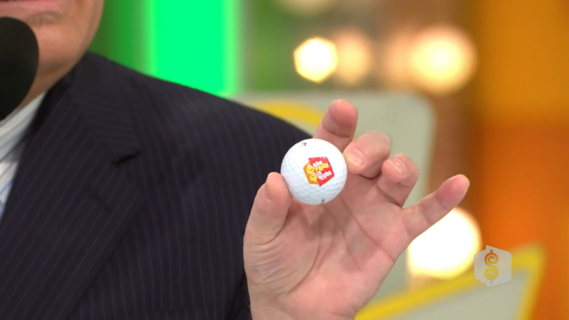 A close-up of a person holding a golf ball with the 'The Price is Right' logo on it, wearing a striped suit.
