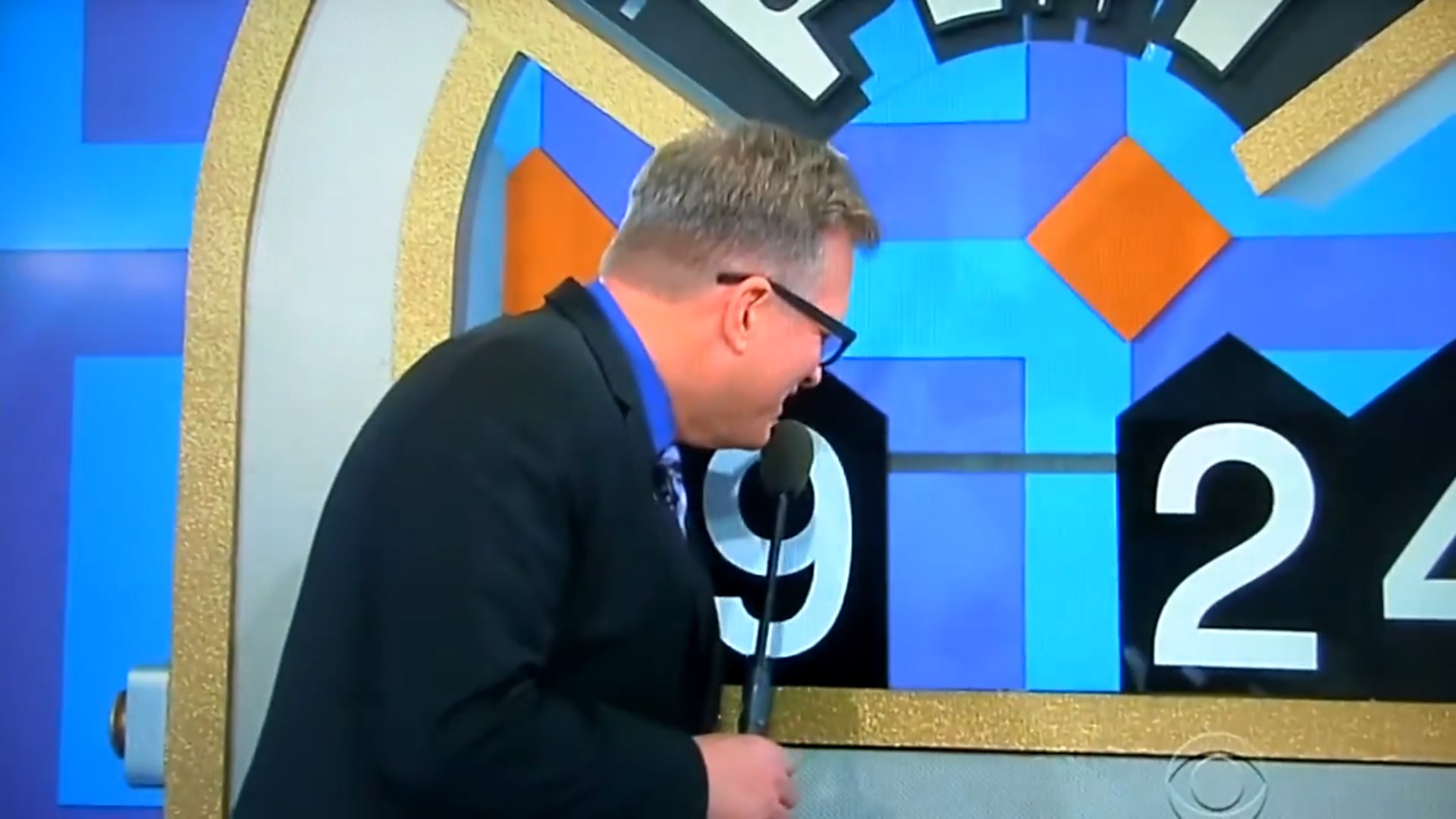 Drew Carey leans in towards a colorful game board displaying the numbers 9, 2, and 4, while holding a microphone.