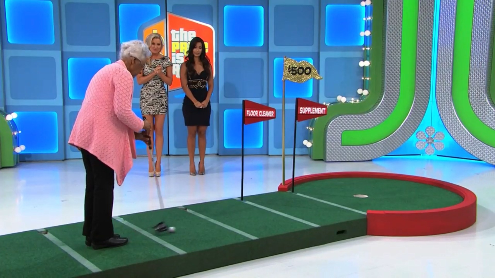 A contestant prepares to putt a golf ball on a putting green with flags indicating grocery product prices, while two female models watch.