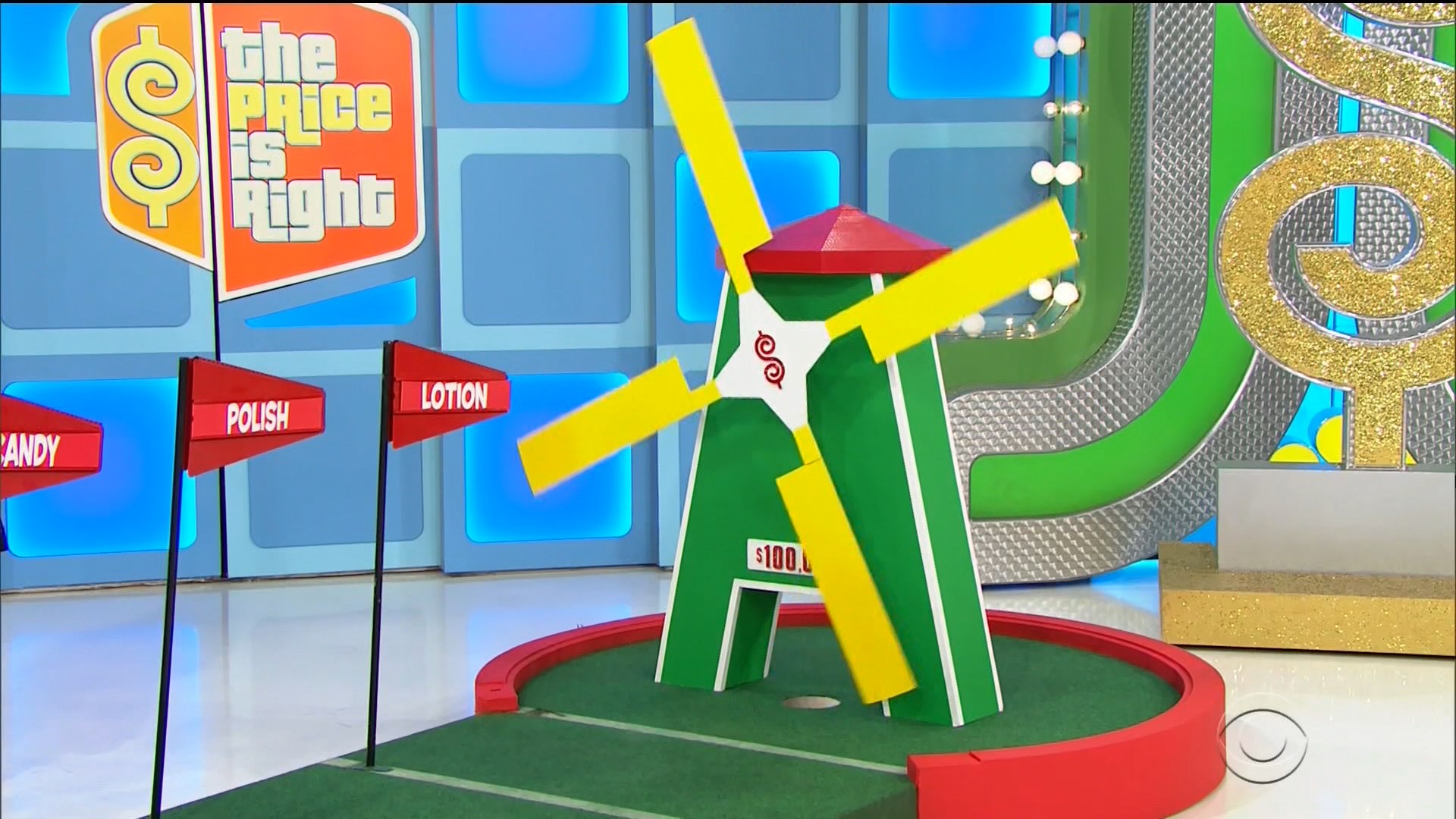 A colorful windmill structure with yellow blades and a sign displaying 0 appears on a green putting green, surrounded by flags for Polish, Lotion, and Candy.