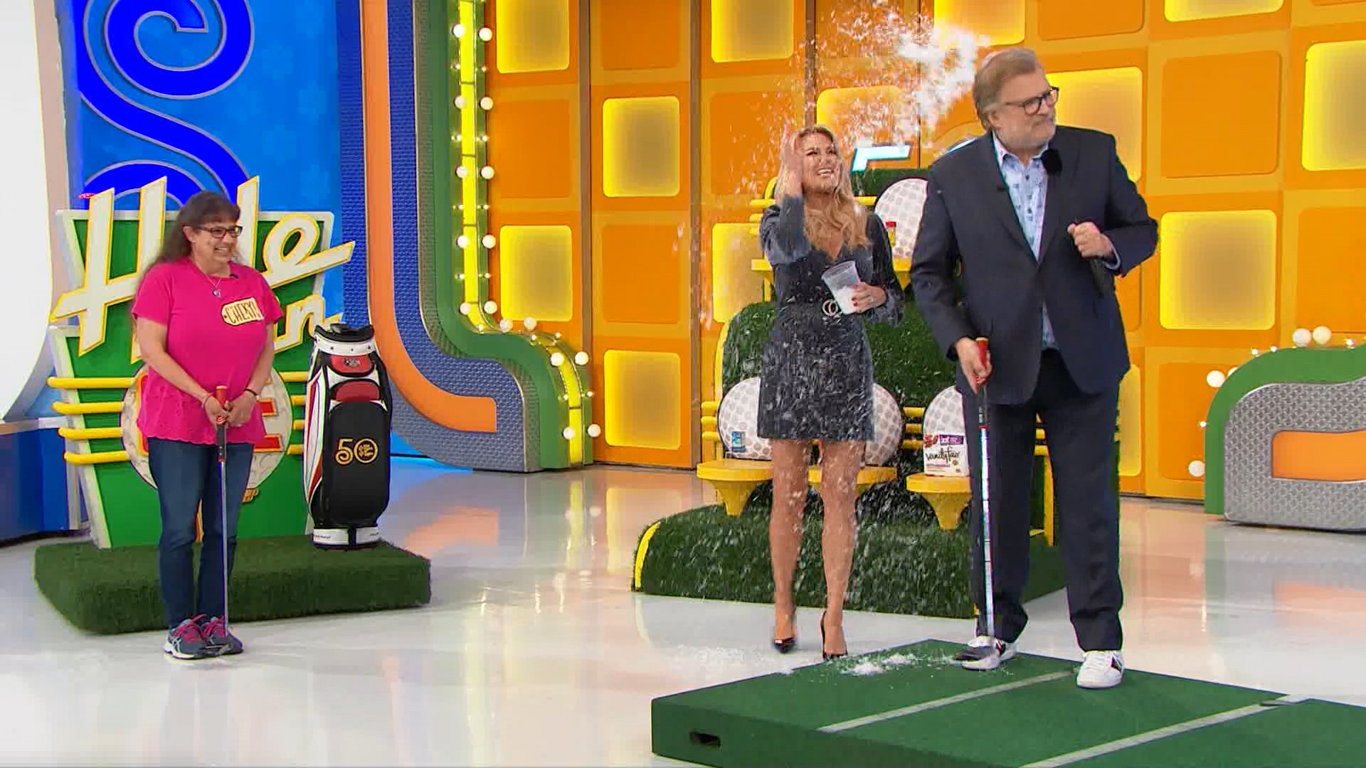 A contestant stands on a putting green with a golf club, while a host prepares to make a putt. The set features the game's logo and elements related to the Hole in One game.