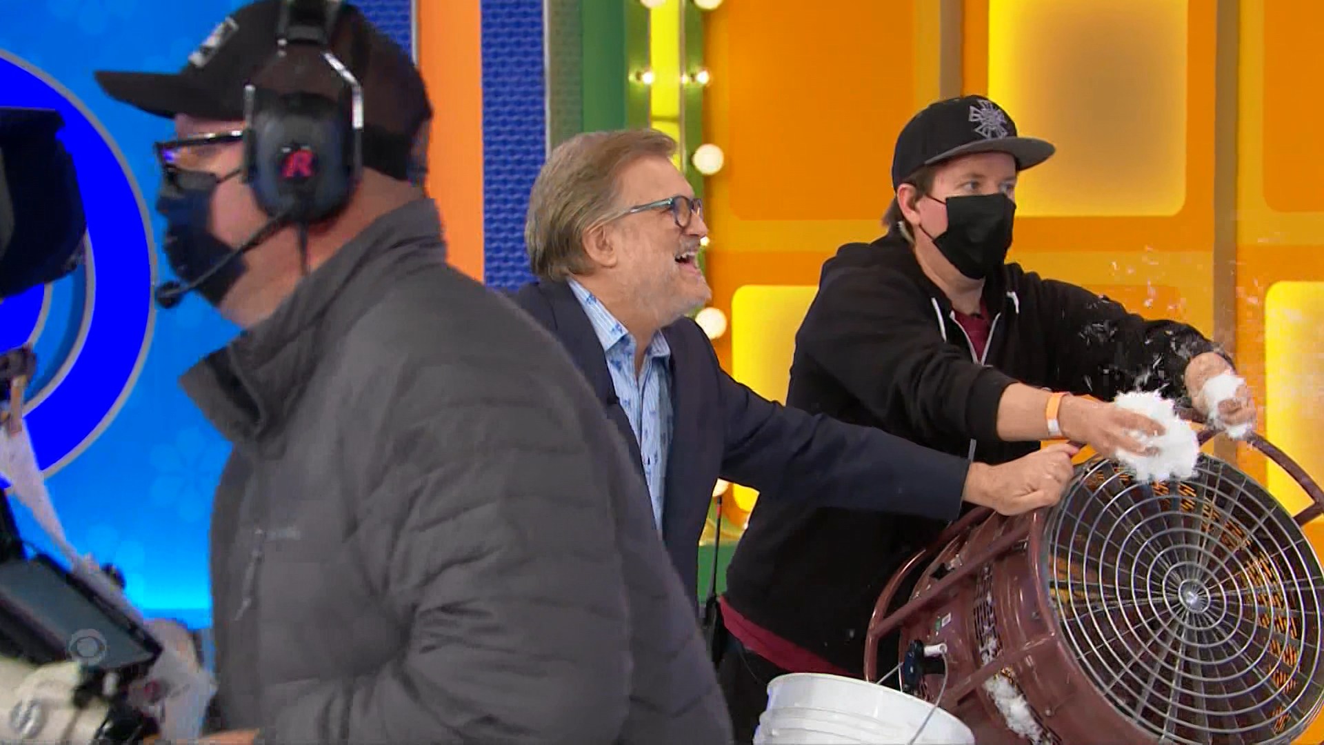 A crew member operates a fan blowing fake snow while another team member prepares it, with a smiling host in a jacket nearby.
