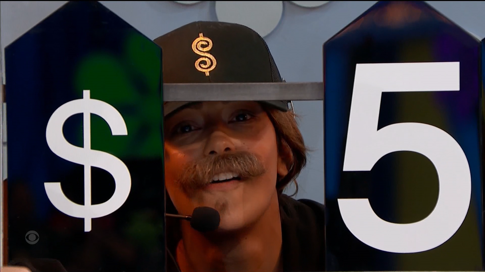 Manuela, wearing a fake moustache, smiling and wearing a hat with a dollar sign, holding a sign showing the number 5 in a game setting.