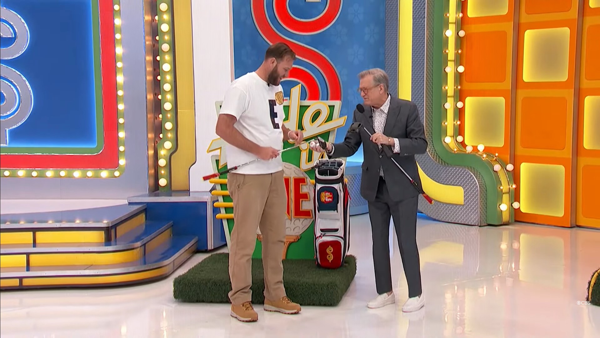 A contestant and the host stand on stage next to a putting green for the game 'Hole in One' on 'The Price is Right'. The contestant holds a golf club while the host shows a golf ball, smiling.