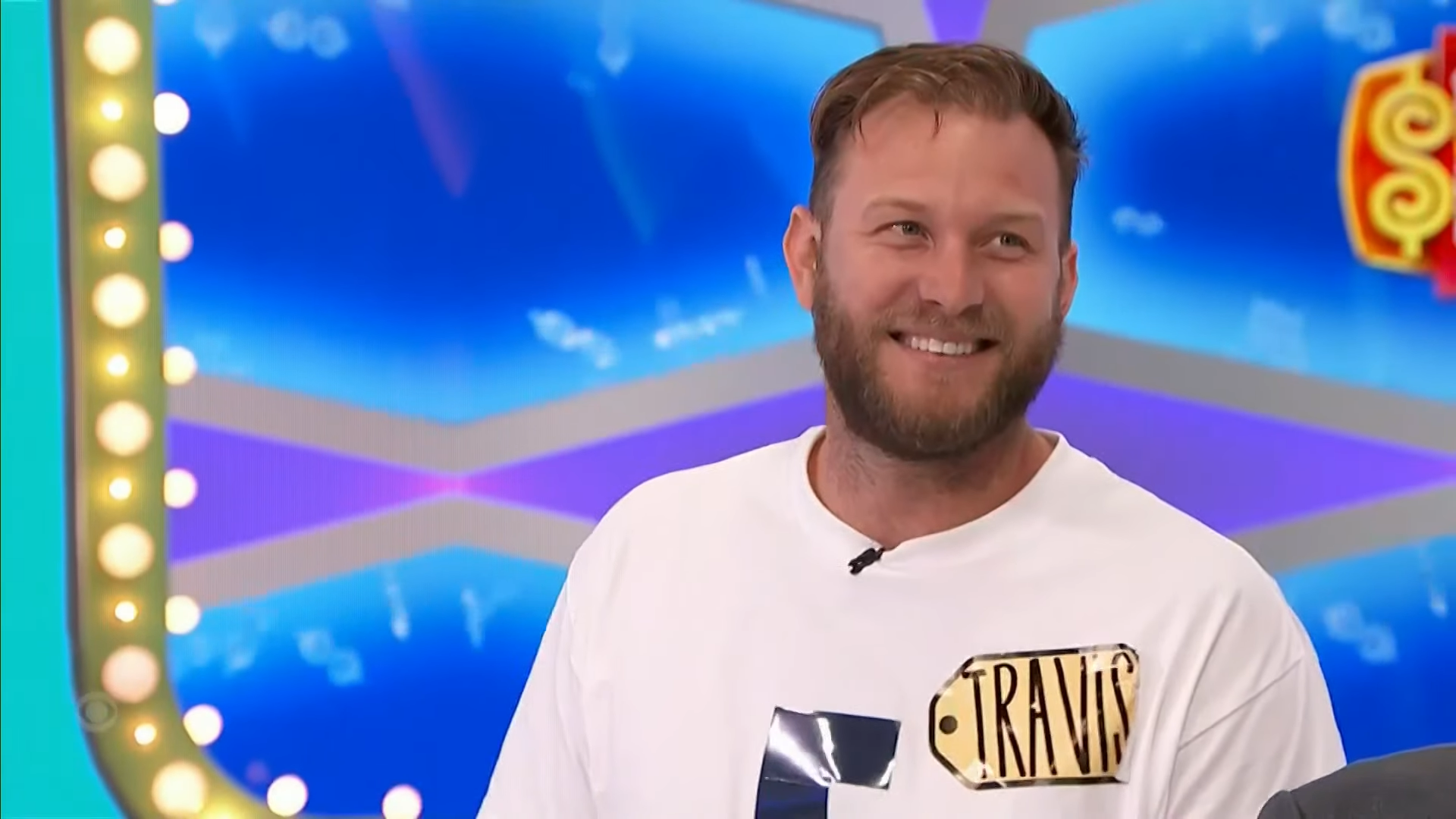 A contestant named Travis smiles while standing in front of a colorful game show backdrop.