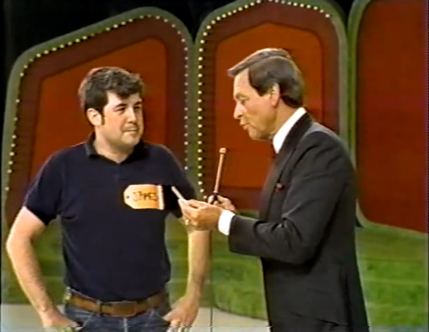 A contestant named James stands on stage during a game show, listening intently to the host, who is holding a pointer and discussing the game details. The stage background features colorful panels.