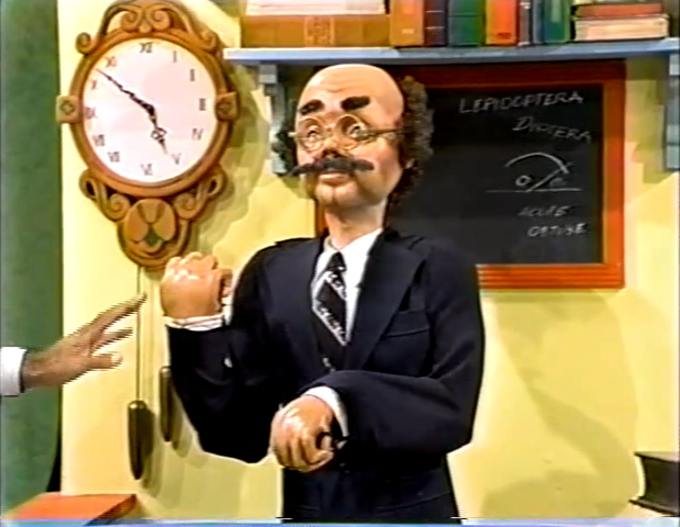 A puppet character resembling a professor in formal attire, complete with glasses and a mustache, is situated in a classroom setting with a clock and a chalkboard behind him.