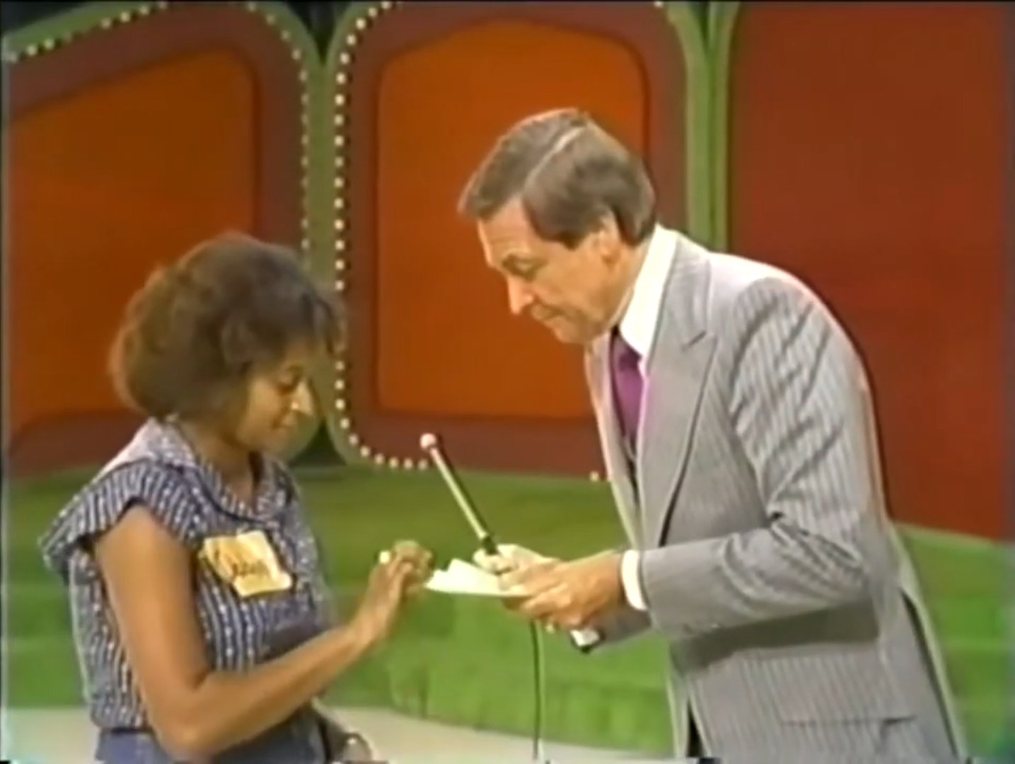 A contestant interacts with a host on stage during a game show, with a colorful backdrop and a microphone in hand.