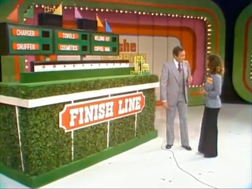 A host and a contestant interact in front of the 'Finish Line' game display, featuring colorful panels and a lush green base.