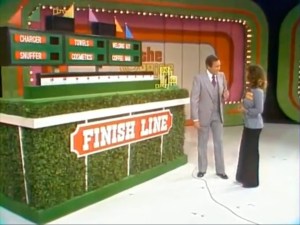 A host and a contestant interact in front of the 'Finish Line' game display, featuring colorful panels and a lush green base.