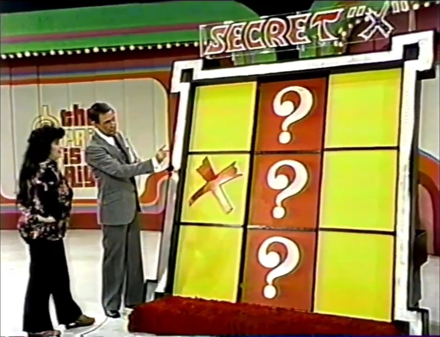 A game board for Secret 'X' from The Price is Right, featuring a Tic-Tac-Toe layout with a visible 'X' in the middle. A contestant and the host are interacting in front of the board.