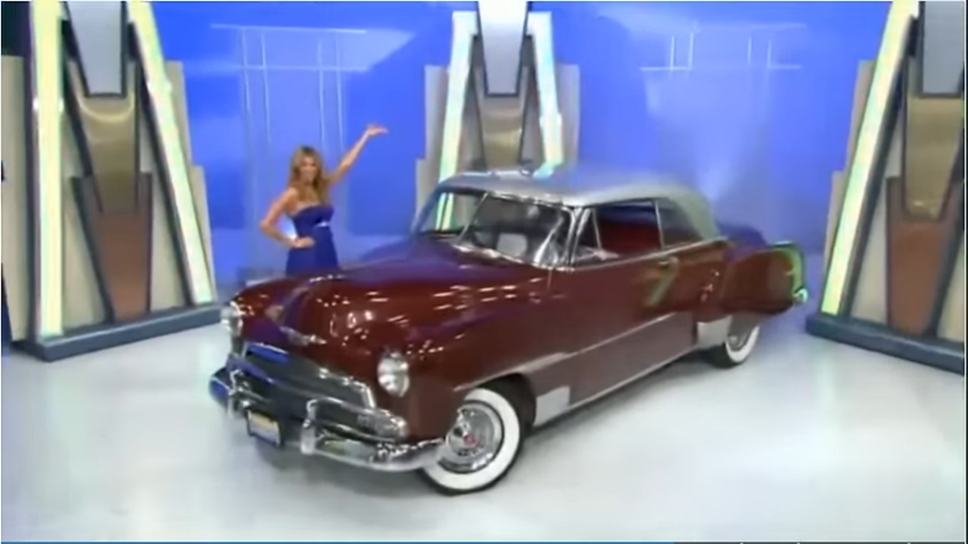 A contestant in a blue dress stands next to a vintage maroon car on a game show set, gesturing towards it.