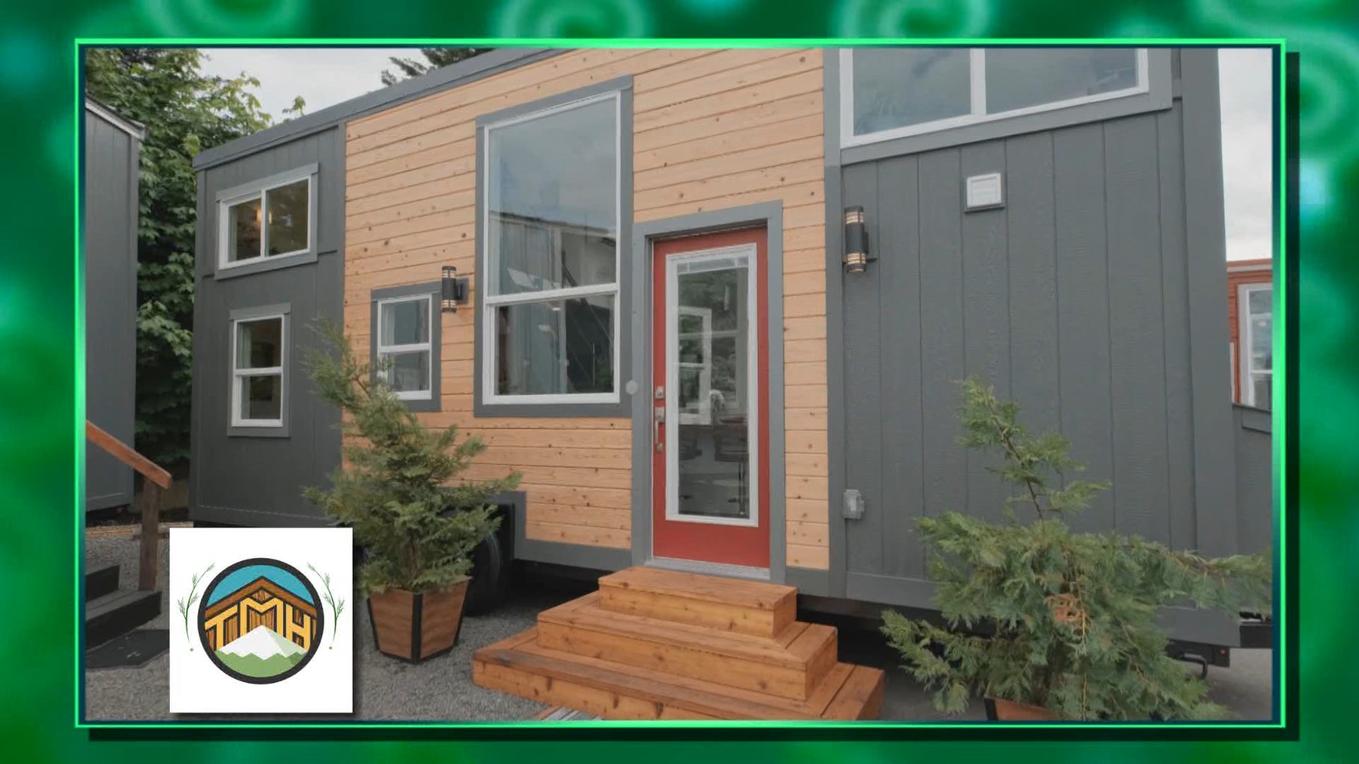 A modern tiny house featuring a combination of wooden and gray paneling, with a front door painted in red and surrounded by potted greenery.