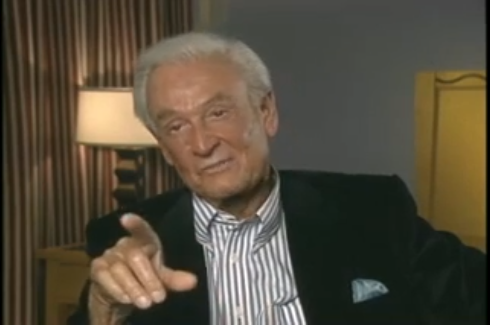 Bob Barker speaking during an interview, gesturing with his hand while wearing a black suit and striped shirt.