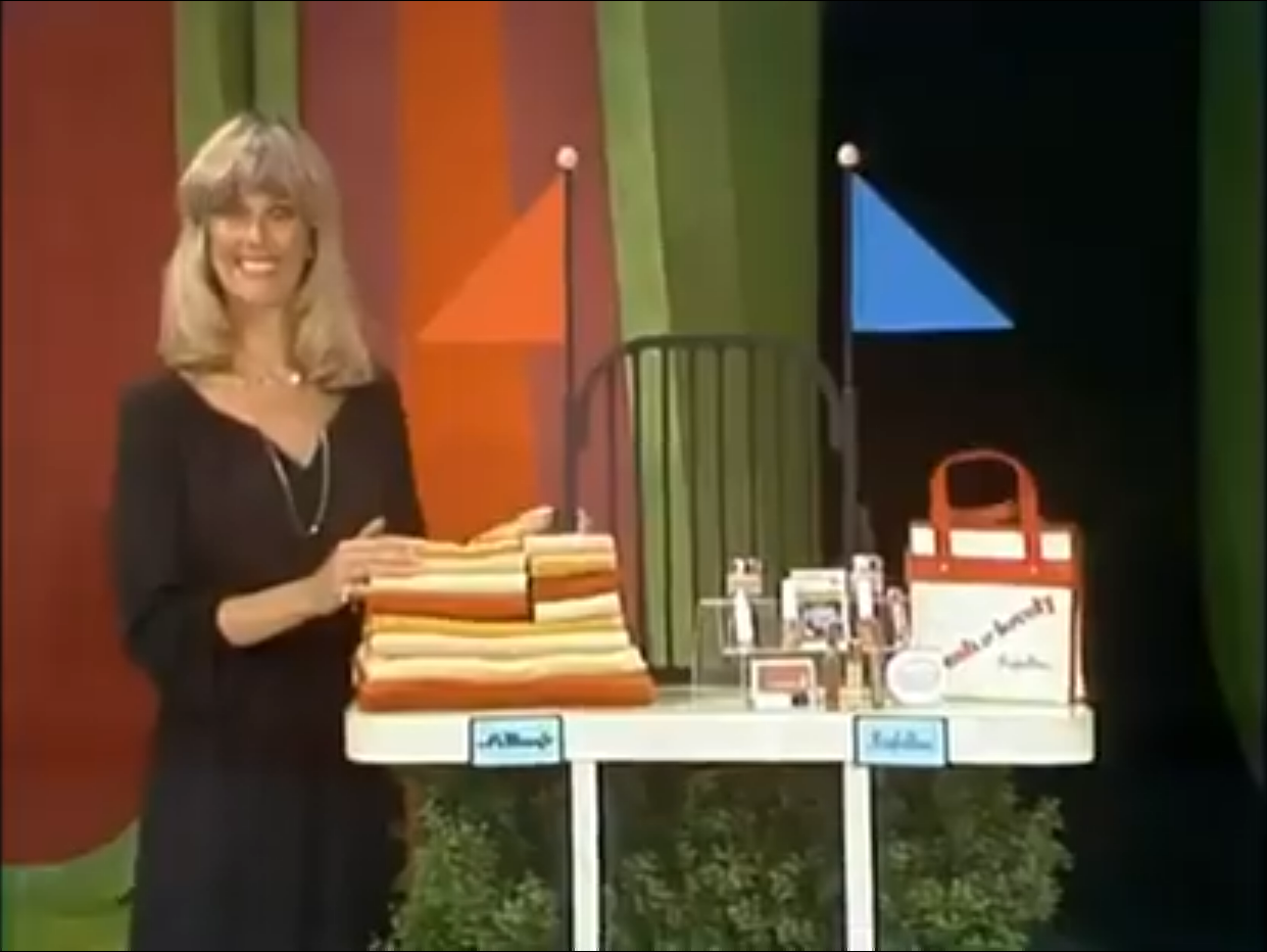 A female contestant stands beside a table displaying a set of colorful towels and various cosmetic products, smiling at the camera during the game 'Finish Line.'
