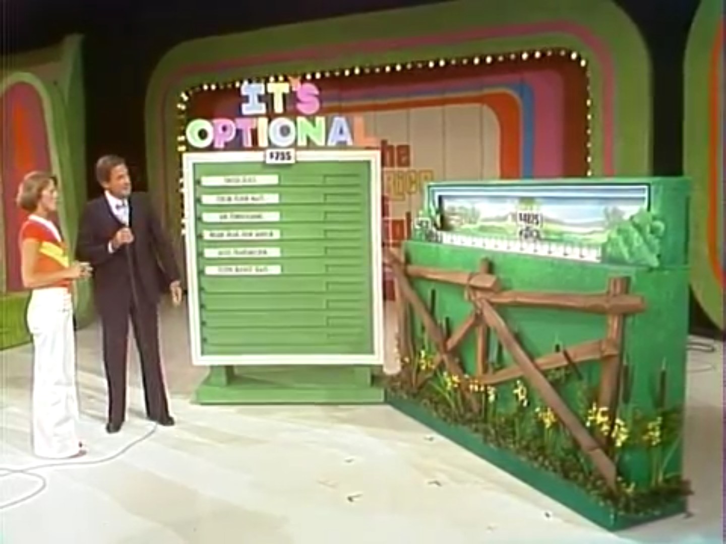 A game show host with a microphone stands next to a female contestant in a colorful outfit, showcasing a large green board with various options for a game called "It's Optional".