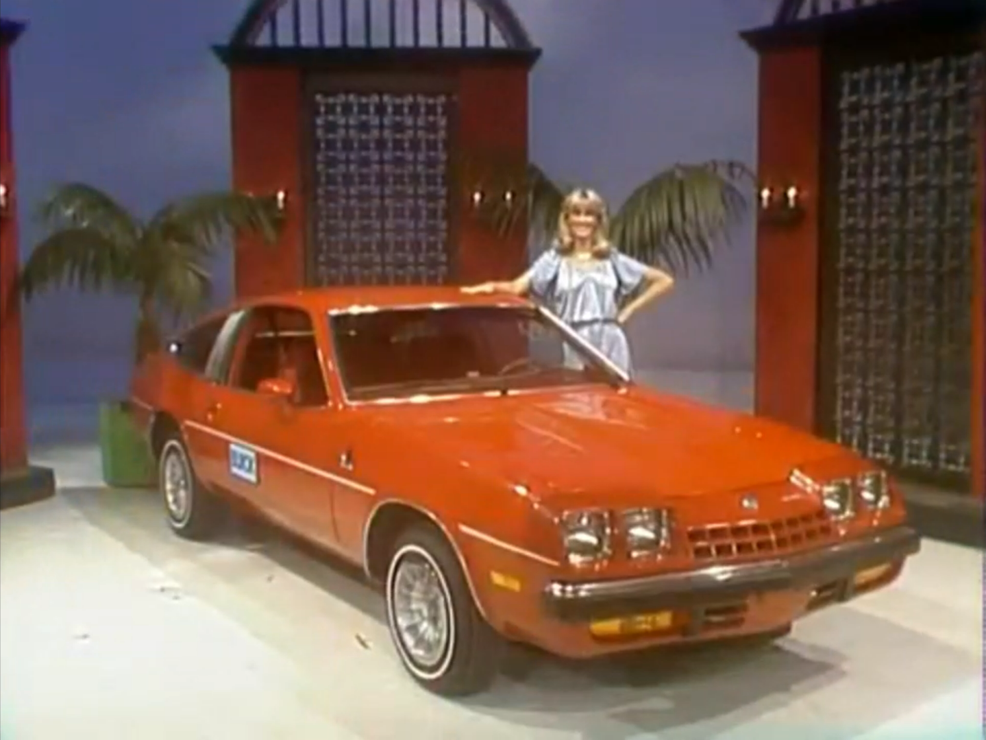 A red 1978 Buick Skyhawk car displayed on stage with a model beside it, set against a colorful backdrop with palm decorations.