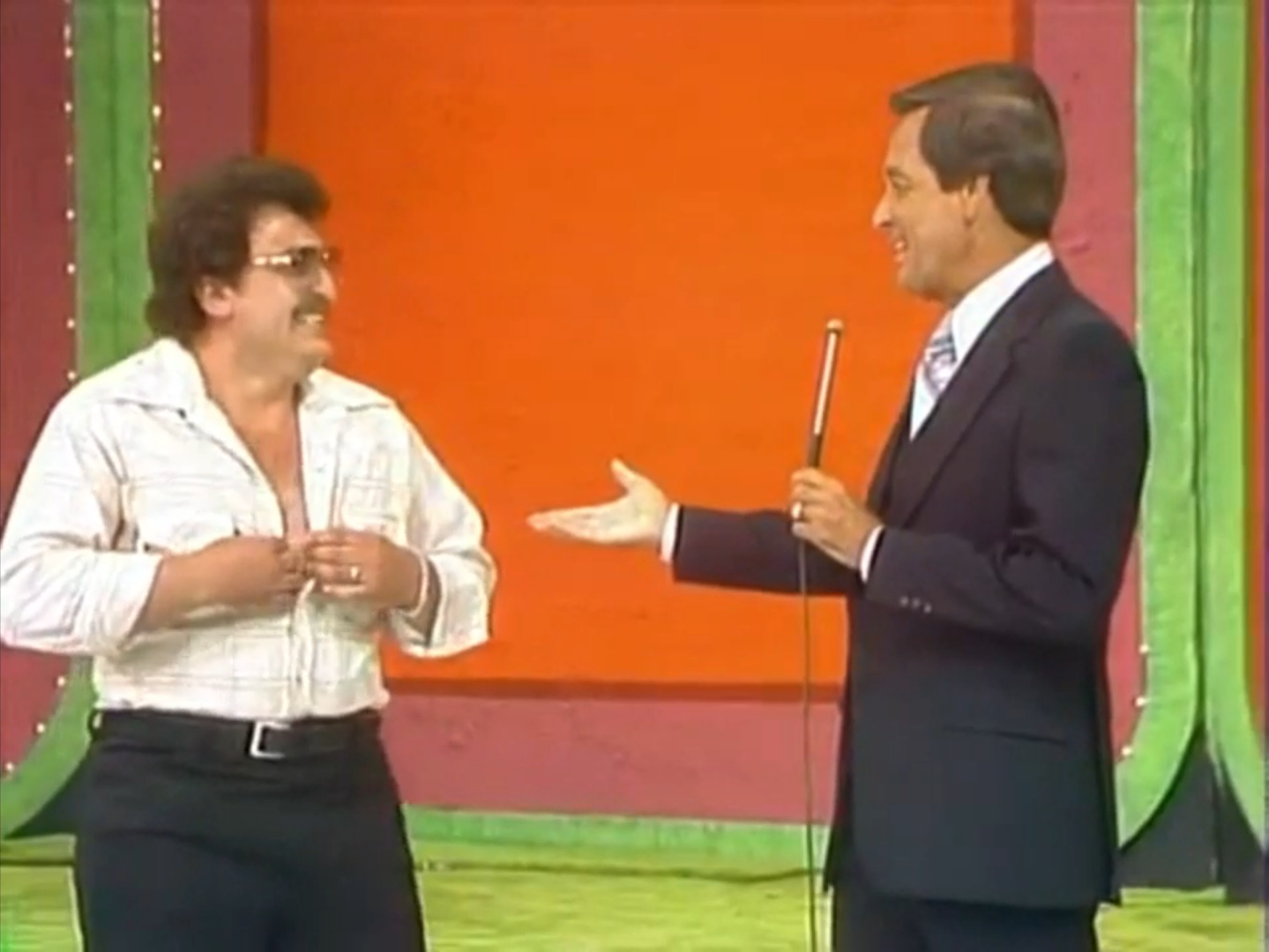 A man in a retro outfit humorously adjusting his shirt while smiling, stands next to a host holding a microphone on a brightly colored stage set.