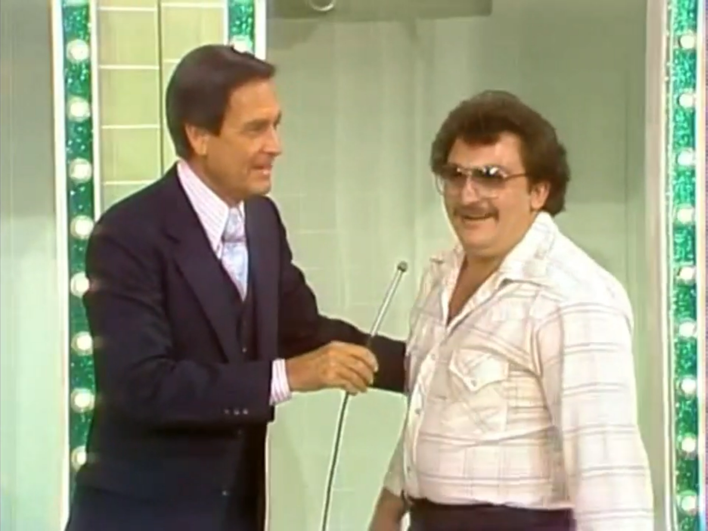 A host and a contestant stand together on the set of a game show, chatting in front of a brightly lit backdrop.