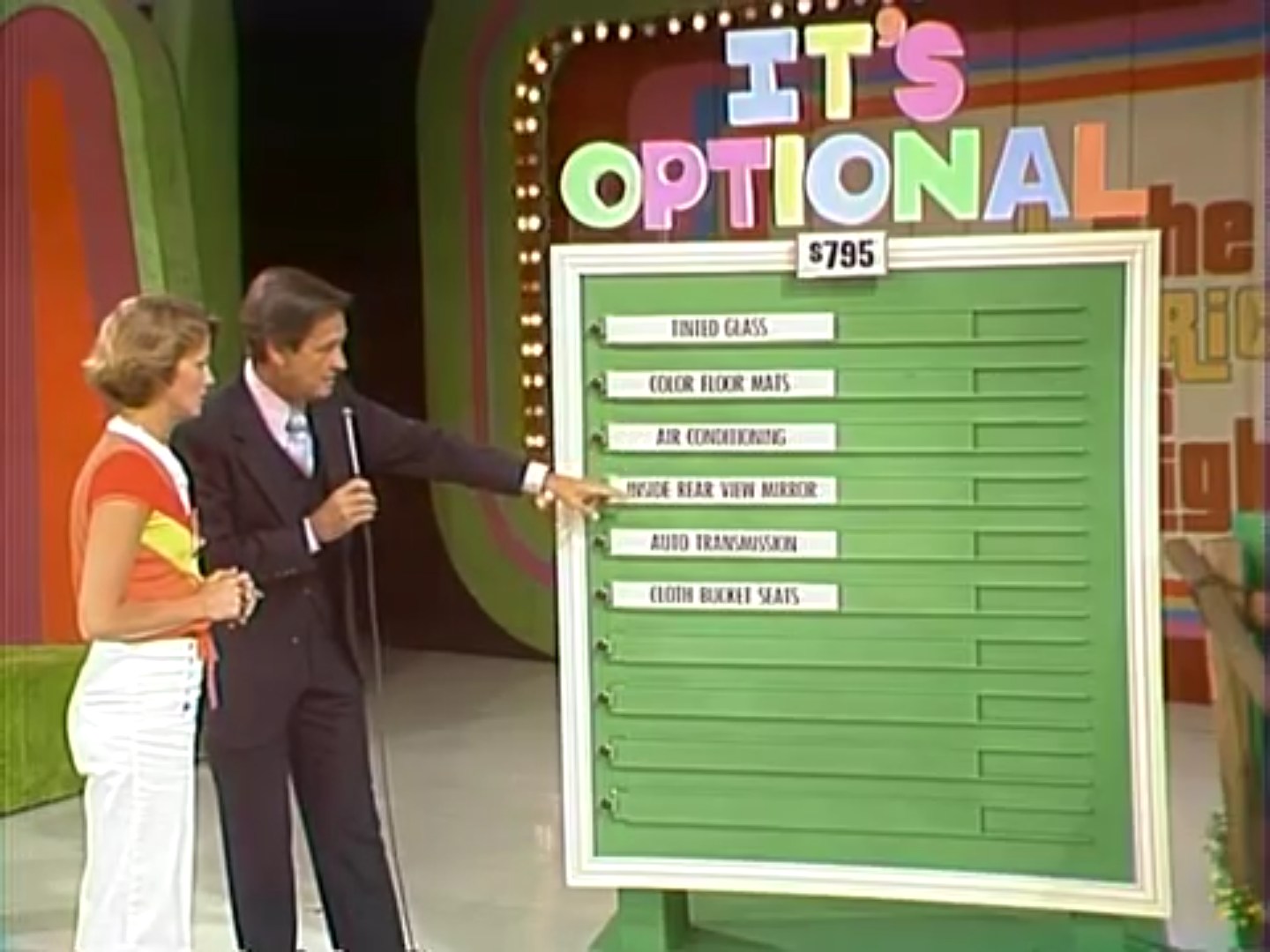 A game show host points to a board with options for a contestant on The Price is Right, displaying prices for features like air conditioning and auto transmission.