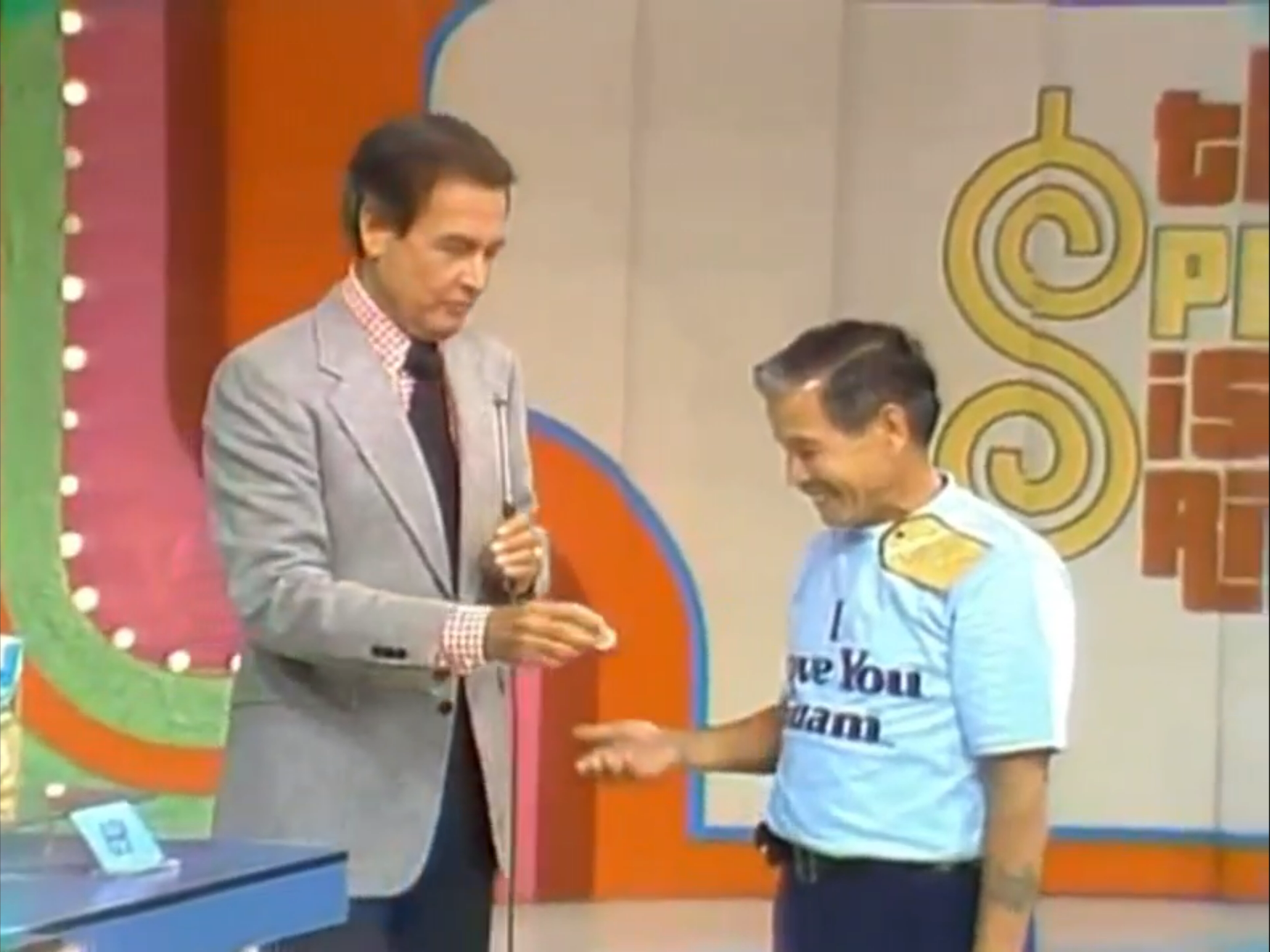 A game show host handing a dime to a contestant on stage, with colorful backdrop and game graphics visible.