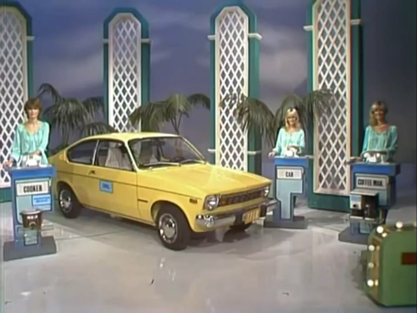 A yellow car is displayed on stage with three women beside it, each at a podium labeled 'COOKER', 'CAR', and 'COFFEE MAKER'.