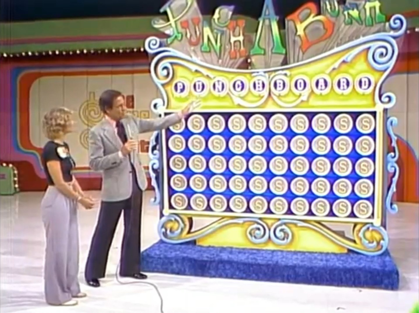A game show host explains the game Punch a Bunch while a contestant watches, standing beside a colorful punchboard featuring various envelopes.