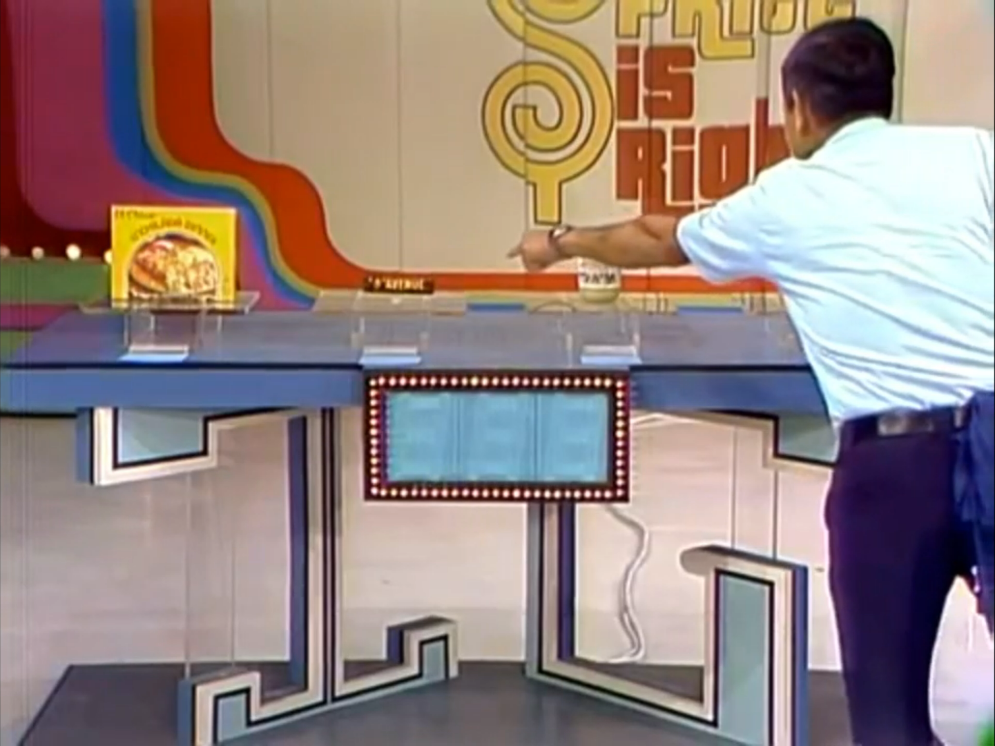 A game show host points at a table featuring displayed grocery products and a logo for 'The Price is Right' in a colorful studio setting.