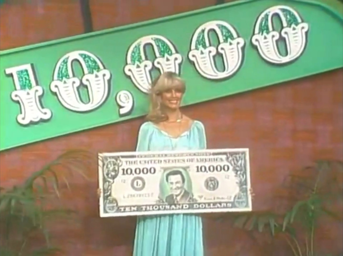 A model on The Price is Right holds a large replica bill for ten thousand dollars, smiling in front of a themed background.
