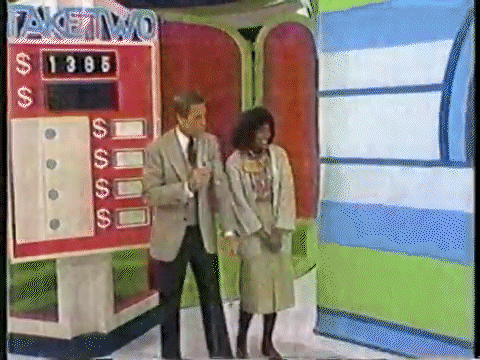 A game show host stands next to a contestant in front of a colorful game board labeled 'Take Two' with a target price displayed.