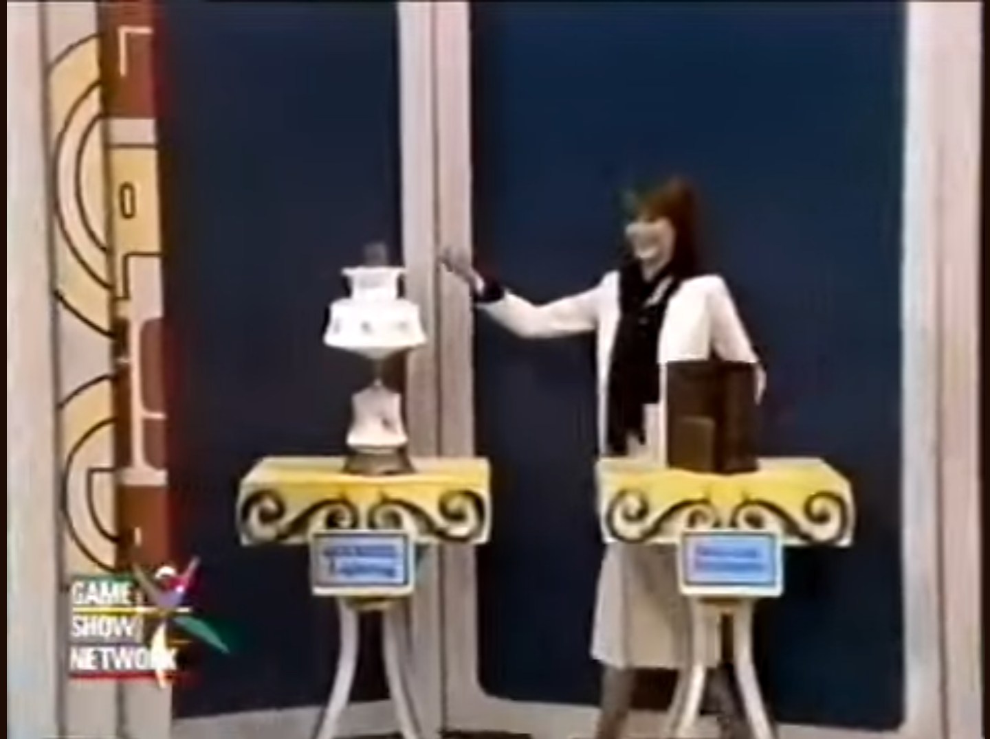 A smiling female game show model presents two small prize podiums on a colorful set. One podium displays a decorative cake, while the other holds a box. The model gestures towards the prizes, engaging with the audience.