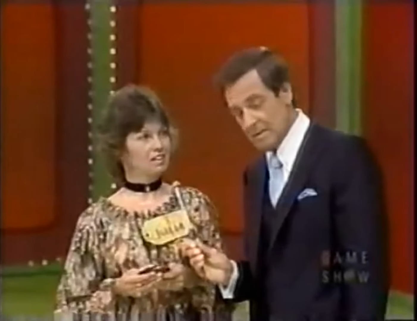 A contestant on a game show looks concerned while holding a prize card, with the host attentively listening to her.