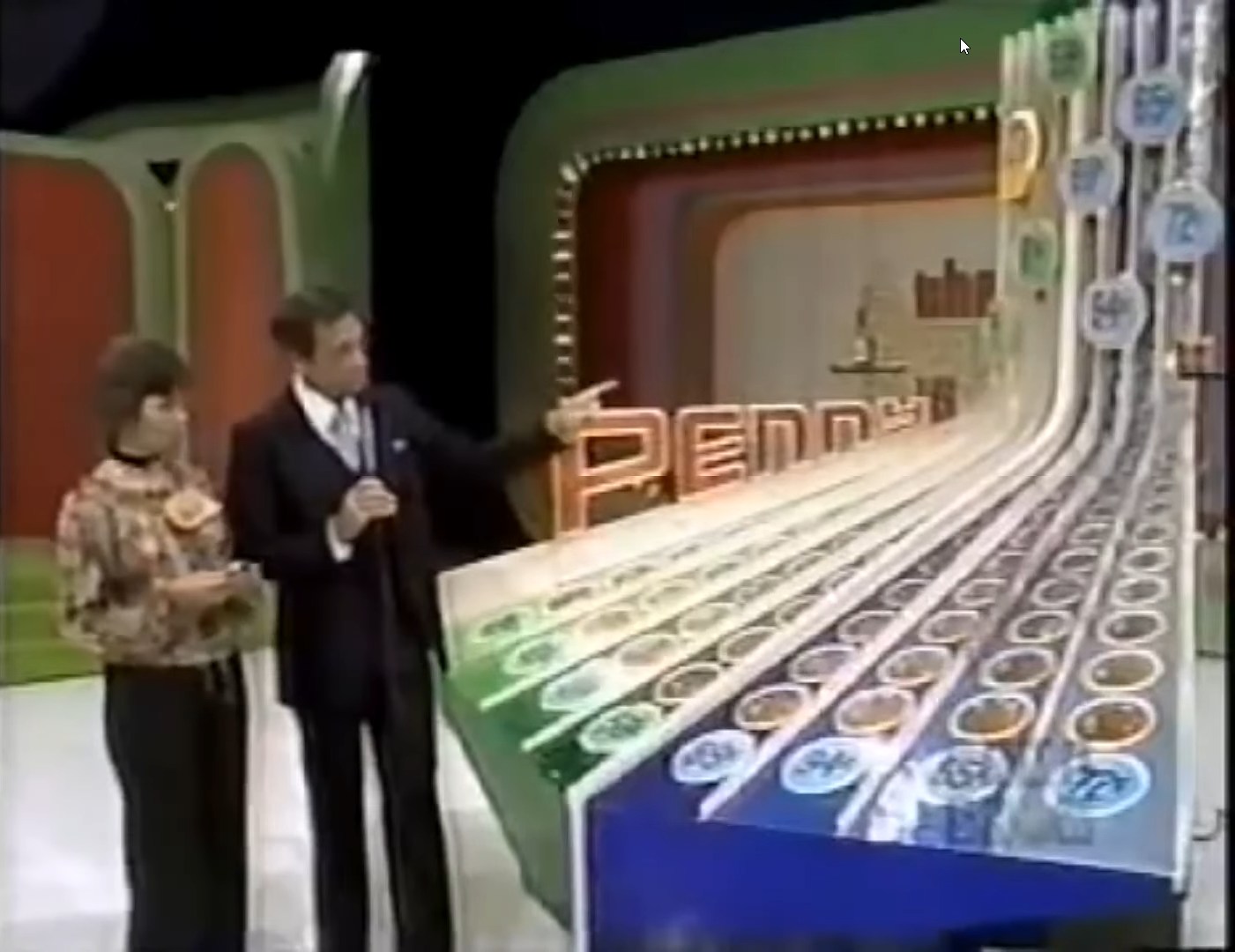 A host points at the Penny Ante game board while a contestant holds a card, showcasing the colorful price display.