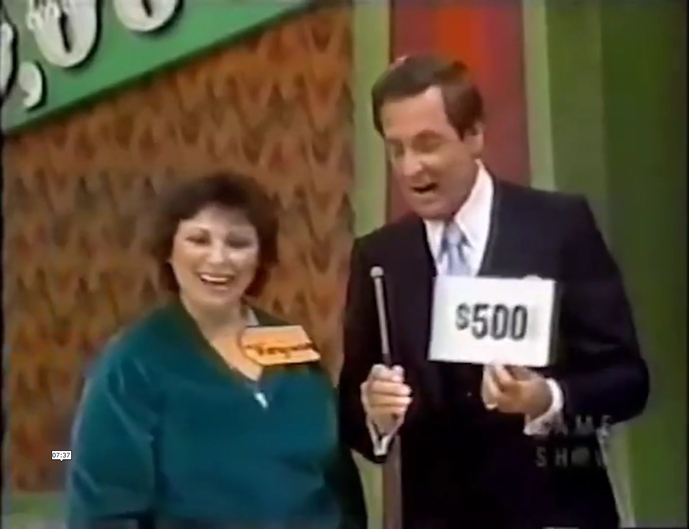 A contestant holding a 0 sign on the set of a game show, smiling alongside the host.