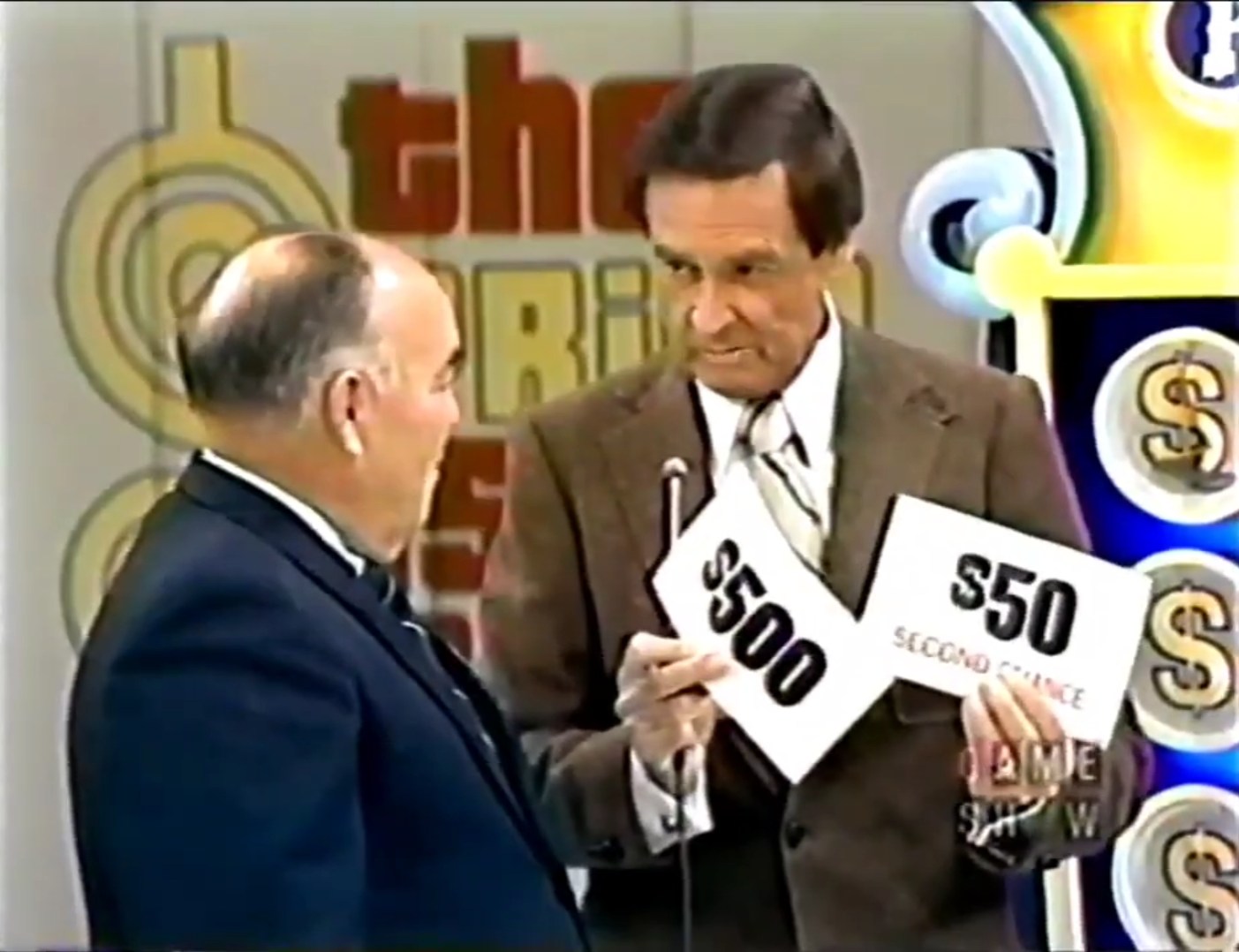 A host holding two prize cards: one shows 0 and the other shows labeled 'Second Chance', with a contestant looking on.