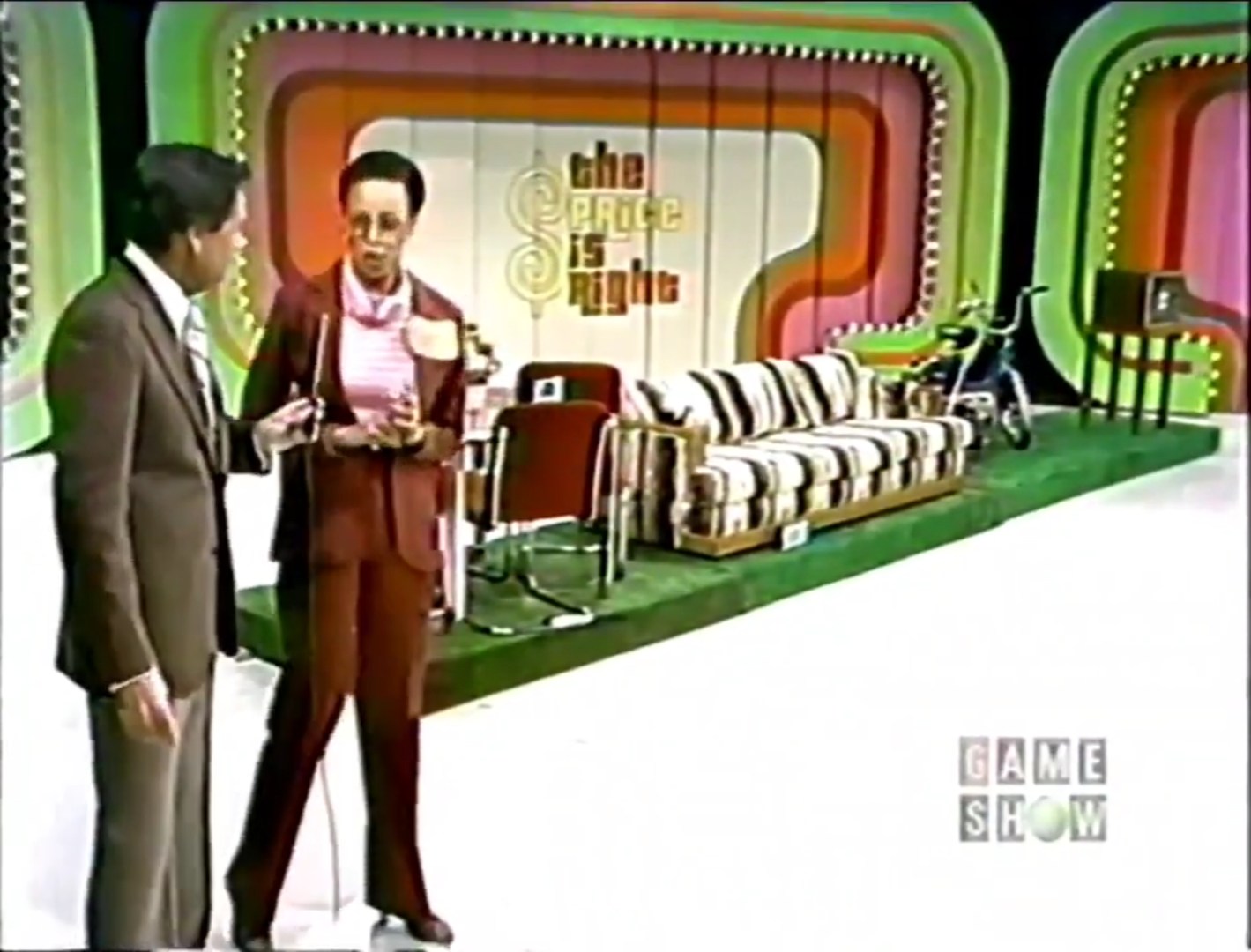 A contestant in a maroon suit is discussing with the host on the set of The Price is Right, featuring a colorful background and various prizes, including a striped couch and a television.