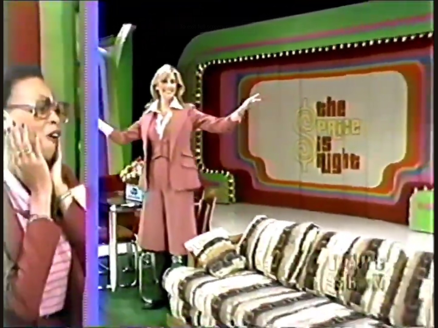 A contestant reacts with excitement while a host gestures in front of 'The Price is Right' set, featuring a colorful backdrop and a patterned sofa.