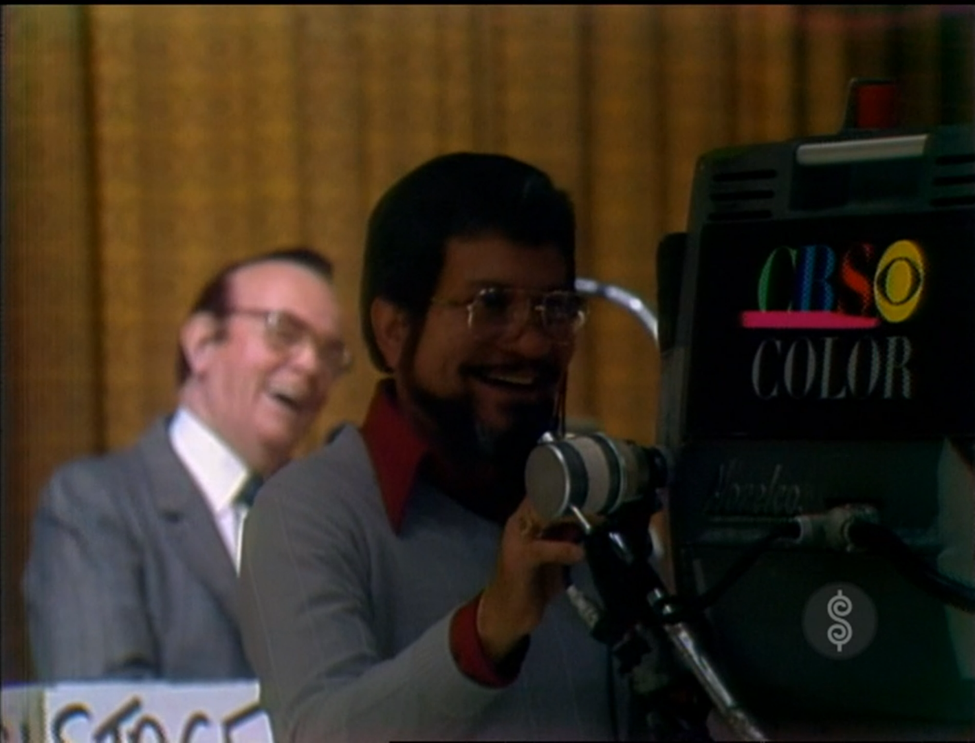 A behind-the-scenes shot from a game show featuring a smiling camera operator in the foreground and a laughing host in the background, with a 'CBS COLOR' logo visible on the camera.