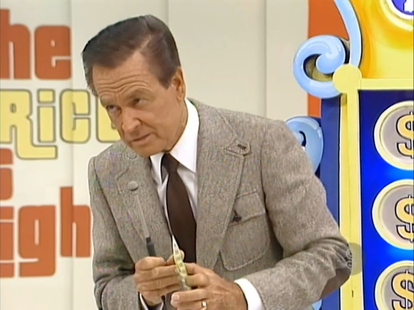 Bob Barker, wearing a grey suit and brown tie, is teasing the contestant by waiting to reveal a slip in Punch a Bunch.
