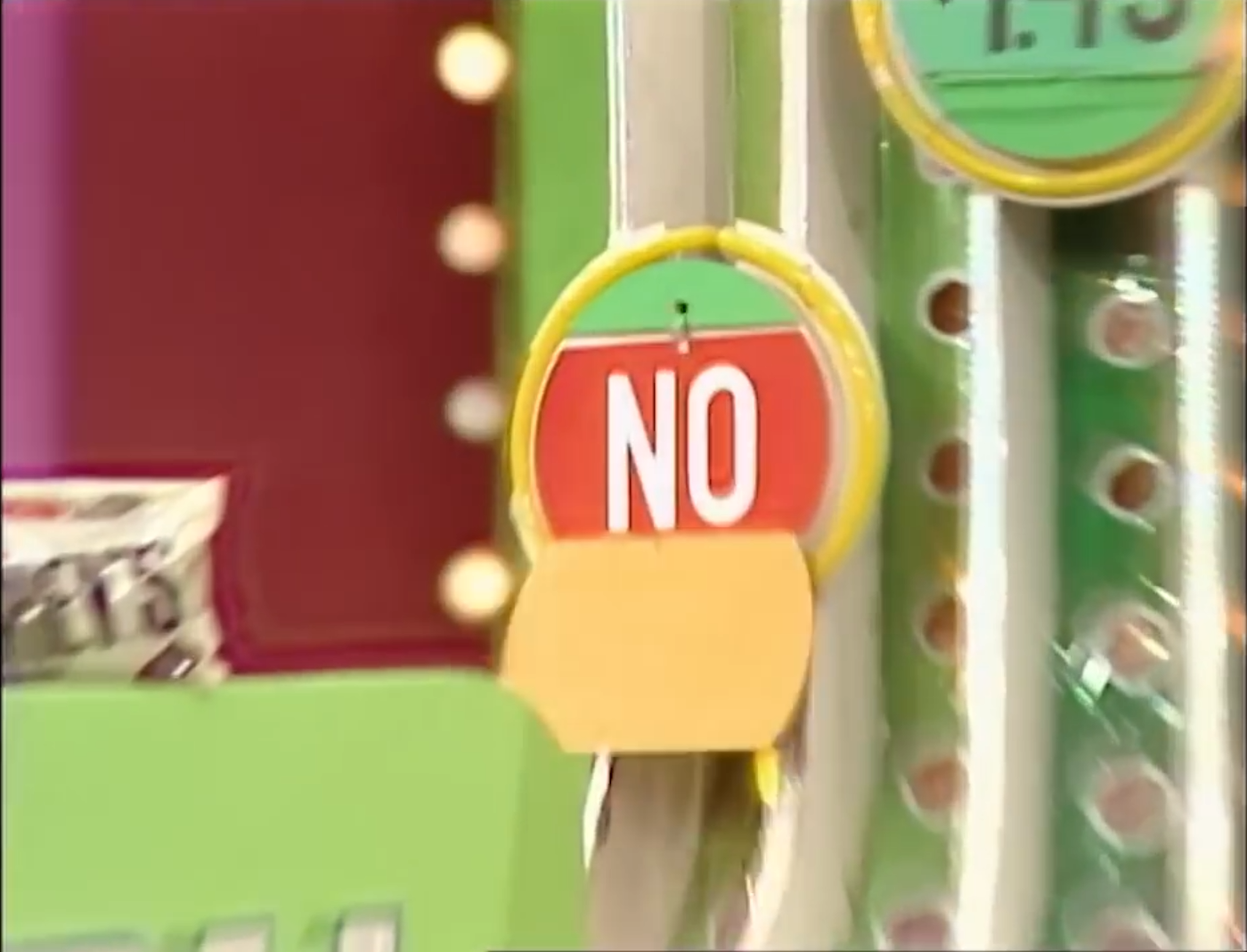 A red and yellow sign displaying the word 'NO' on a game show set, with green price tags and colorful game elements in the background.
