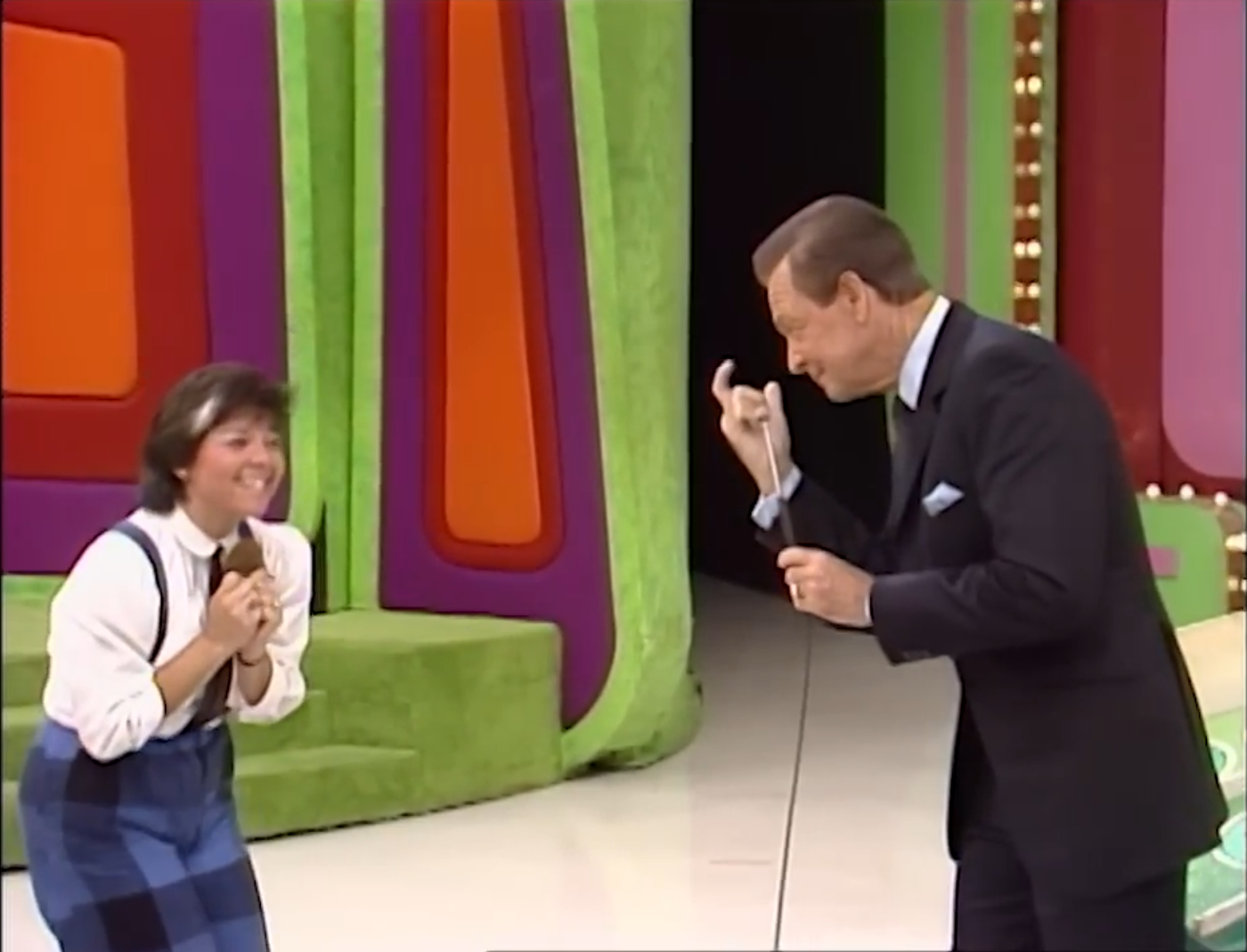 A contestant smiles excitedly while holding a microphone, as a game show host gestures enthusiastically in front of a colorful game set.