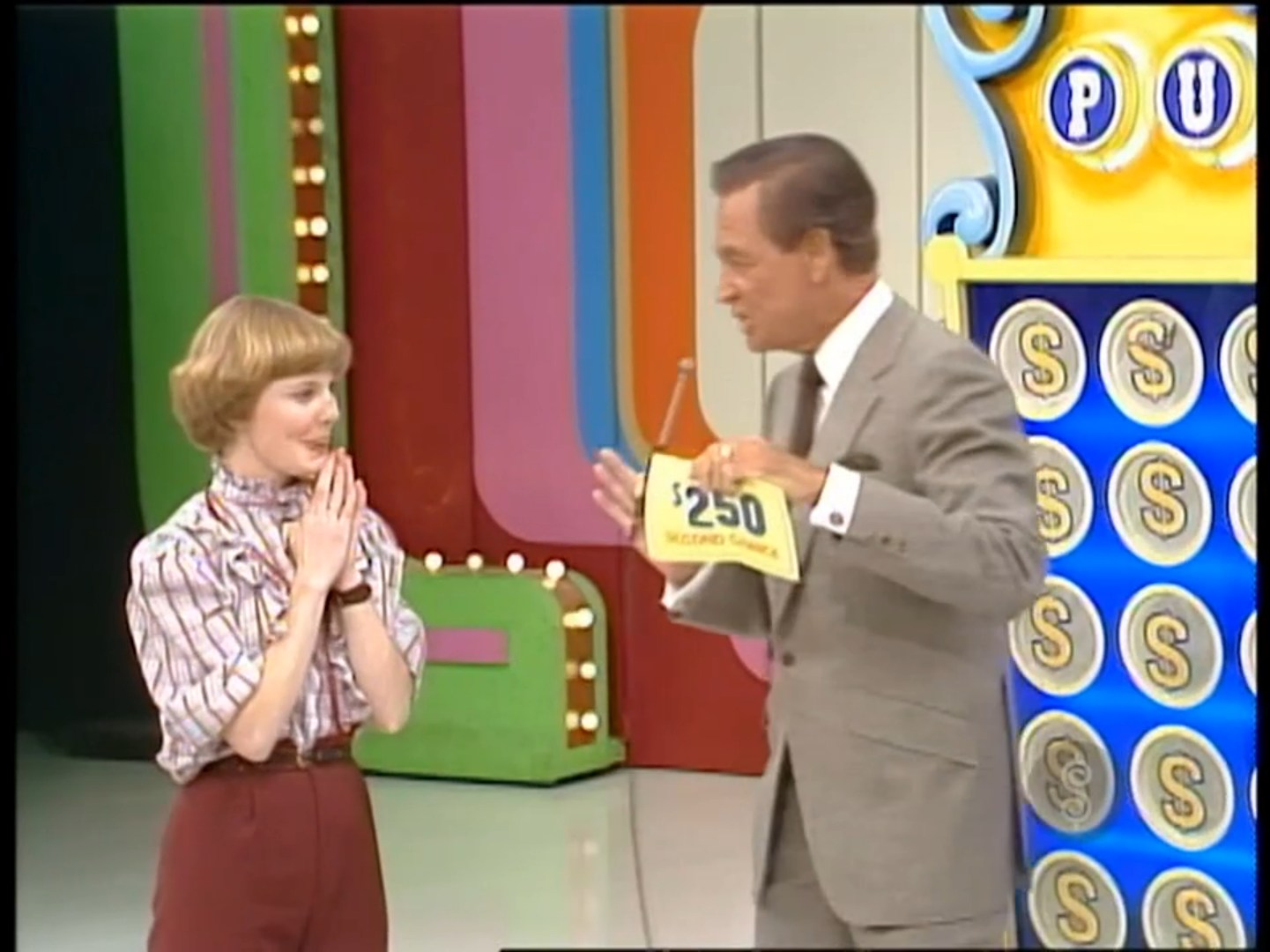 A contestant, visibly excited, stands with clasped hands while a host presents a 0 prize on The Price is Right set, featuring colorful backgrounds.