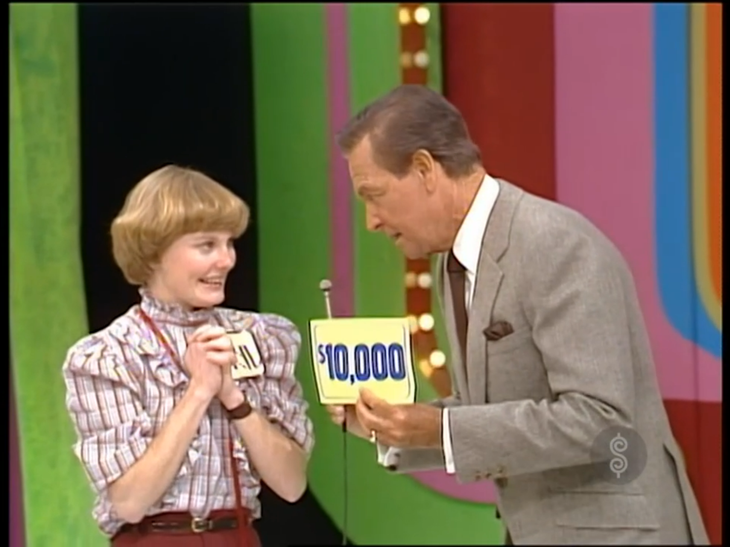 A contestant displays a sign reading ',000' while showing excitement, accompanied by a host who leans in to engage with her. The background features vibrant colors typical of a game show setting.